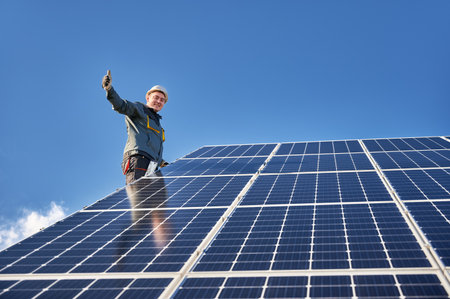 Male worker giving thumbs up and smiling while mounting blue photovoltaic solar panel under beautiful cloudy sky. Concept of alternative energy sources and innovations.の写真素材