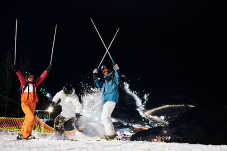 Friends skiers in winter jackets and helmets having fun at ski resort in the mountains in winter, skiing at night, illuminated ski pistes are downhill on background. Concept of healthy lifestyleの写真素材