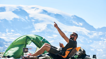 Male tourist sitting in chair near tent in empty rocky alpine mountains, using smartphone in sunny...