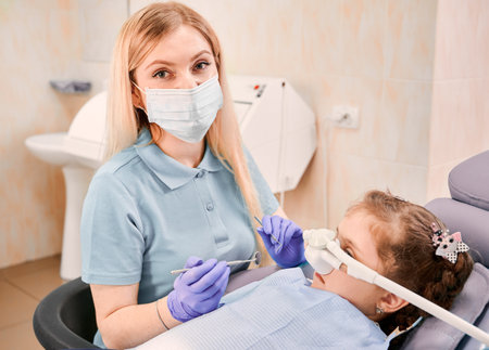 Close up dentist in medical face mask holding dental instruments while girl lying in dental chair with inhalation sedation at dental office. Concept of pediatric, sedation dentistry and dental care.の写真素材