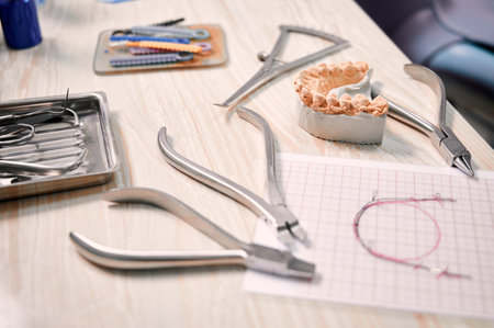 Desk with orthodontic tools, ligature ties, dental teeth model and wire braces drawing. Ligature wire cutters, pliers, multicolored teeth correctors and lower jaw model on table in dental office.の写真素材