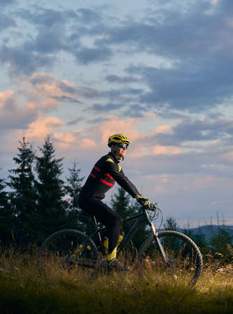 Young man cyclist riding bicycle on sunset in the mountains. Bicyclist wearing helmet, sports glasses and uniform. Side view. Evening sky on background. Concept of active lifestyleの写真素材