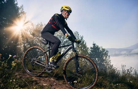 Young sportsman riding his bicycle in the mountains in early foggy morning going downhill. Sun is...