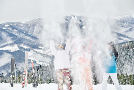 Three skiers in ski suits throwing fresh powder snow high in the air, having fun at ski resort with beautiful mountains on background. Concept of winter sport activities, fun and relationships.の写真素材