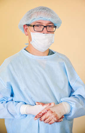 Portrait of male doctor in glasses looking at camera. Man surgeon wearing sterile gloves, blue surgical uniform, medical cap, protective face mask. Concept of medicine, healthcare workers and surgery.の写真素材