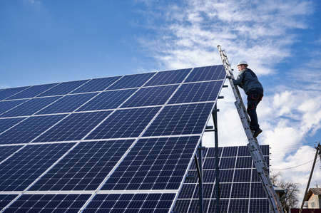 Side view of man technician in safety helmet climbing the ladder to get to the top of photovoltaic solar module. Male worker mounting stand-alone solar panel system. Concept of alternative energy.の写真素材
