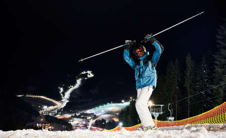 Skier keeping his hands up, standing on hill getting ready for a night ride. Beautiful illuminated ski pistes are down on background in night time. Concept of extreme sportの写真素材