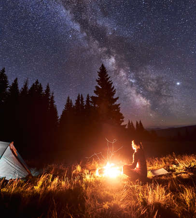 Side view of female tourist having a rest by campfire on background of tent and spruce forest under starry sky on which milky way is visible. Bonfire with a big flame under the night skyの写真素材