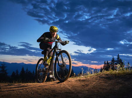 Back view of young man cycling bicycle under beautiful night sky. Male bicyclist in safety helmet riding on hillside road under blue cloudy sky in the evening. Concept of sport and active leisure.の写真素材