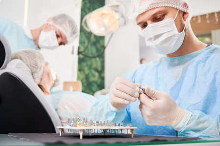 Dentist on foreground, wearing disposable sterile clothes, putting surgical nozzle on drill before...