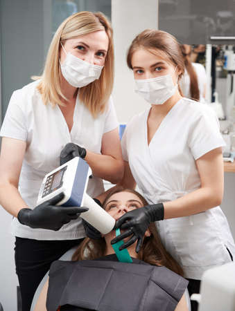 Doctors using portable dental x-ray machine while examining female patient teeth in dental office. Young woman lying in chair while dentists performing intraoral scanning with modern equipment.の写真素材