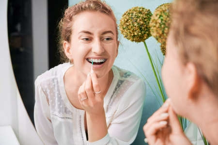 Portrait of a beautiful woman in dentists office demonstrating interdental brushing, looking in huge mirror, wearing dental ceramic and metal brackets. Concept of dentistry and orthodontic treatment.の写真素材