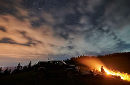 Wide angle view on cloudy starry night in the mountains and a man enjoying nature, sitting on logs...
