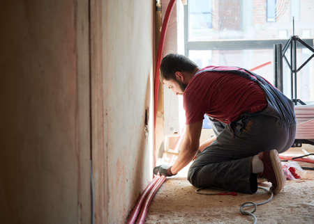 Young plumber connecting water radiator to heating system in new unfinished apartment. Crouching man...