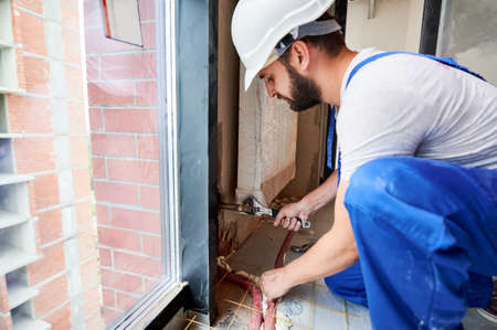 Side view of bearded young man in work overalls installing heating radiator in room. Male worker in...