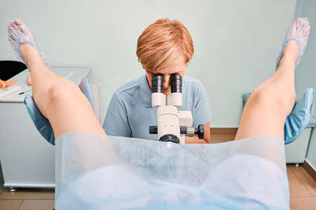 Front view of doctor in blue shirt examining woman with colposcope in gynecological clinic. Female patient laying in gynecological chair while gynecologist doing colposcopy examination.の写真素材