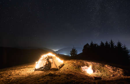 Fantastic view of starry sky over mountain valley with tourist tent and male traveler guitarist. Young man hiker sitting inside illuminated camp tent and playing guitar under night sky with stars.の写真素材