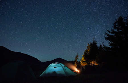 Beautiful view of night starry sky over grassy hill with tourist tents and trees. Mountain valley with illuminated camp tent under night sky with stars. Concept of hiking, night camping and astronomy.の写真素材