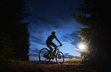 Back view of man cyclist riding bike under blue evening sky with clouds. Silhouette of bicyclist riding bicycle on the trail in night mountain forest. Concept of sport, biking and active leisure.の写真素材