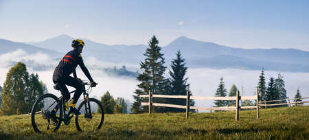 Man riding bicycle on grassy hill and looking at beautiful misty mountains. Male bicyclist enjoying panoramic view of majestic mountains during bicycle ride. Concept of sport, bicycling and nature.の写真素材