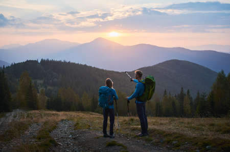 Athletic man with tourist backpack holding up trekking stick in his hand, pointing to sun rising...