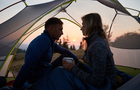 Silhouette of family pair, who relaxing in tent early morning after cold mountain night on background of pink shadows sky during sunrise. Mountain recreation in camp.の写真素材
