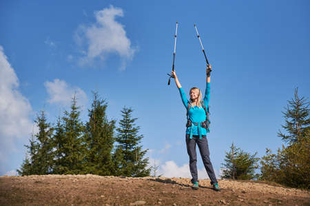 Happy blonde triumphantly raising her hands holding trekking sticks after she climbed on stone top of hill, on the background of several green trees and beautiful blue sky.の写真素材
