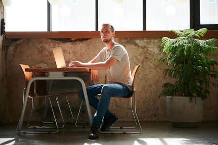 Young handsome man with beard sitting on chair and working or studying in laptop at home or cafe. Concept of process pensive working.の写真素材