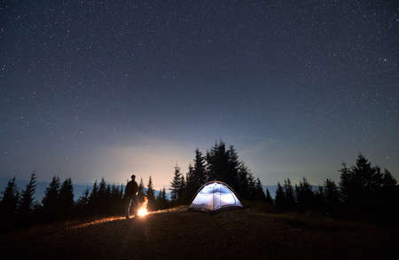 View of night starry sky over mountain valley with male hiker, illuminated camp tent and campfire....