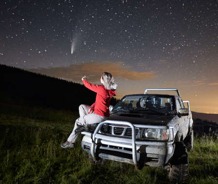 Young woman in the mountains watching beautiful starry night and a comet, sitting on car hood. Illuminated foreground. Bright skyline on background. Astrophotography conceptの写真素材