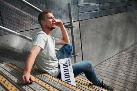 Side view of male with piano keyboard is sitting on stairs. Young man in grey T-shirt is resting on...