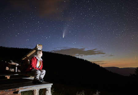 Side view of woman sitting on wooden bench in mountains and watching starry sky. Female in red jacket is relaxing near forest and looking on comet Neowise. Concept of resting in mountains.の写真素材