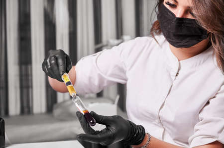 Close up of young woman scientist in medical mask holding test tube with blood sample. Female technician in sterile gloves using syringe and preparing material for plasmolifting process.の写真素材