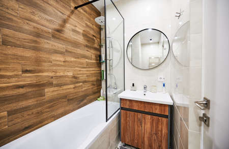 Stylish and comfortable version of bathroom design in small space. White color of rectangular bathtub and wash basin in harmonious combination with round mirror supplemented by wooden decor.の写真素材
