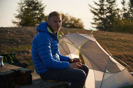 Male traveler holding cup of coffee and smiling while sitting on wooden bench near camp tent. Man hiker with mug of hot drink in his hand resting outdoors. Concept of hiking, travelling and camping.の写真素材
