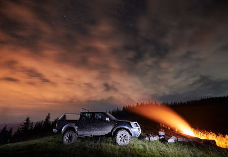 Fantastic scenery of night starry sky over grassy hill with car, wooden planks and campfire....