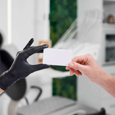 Cropped closeup snapshot of dentist hand in black glove giving business card to patients hand. Dental office on blurred background. Selective focus. Concept of dental advertisementの写真素材