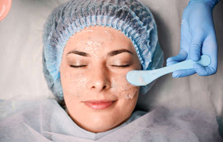 Close up of beautician hand applying cosmetic mask on young woman face. Female client in disposable shower hat having skincare procedure in beauty salon. Concept of skincare and facial treatment.の写真素材