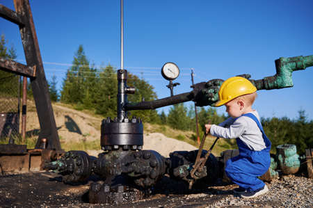 Adorable little boy in construction helmet using industrial wrench while repairing pipe of oil well pump jack. Cute kid oil worker tightening bolt on pipe while working in oil field.の写真素材