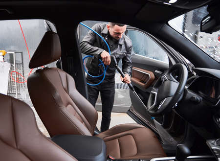 View from inside a car on young man washing car on carwash station outdoor. Handsome driver cleaning automobile, using high pressure water.