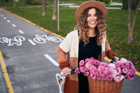Cheerful pretty girl with gentle smile riding on bicycle with bouquet of flowers in basket. On the background bike paths and green lawns.の写真素材