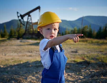 Cute child oil well worker looking at camera and pointing finger at something while standing in oil field. Adorable child wearing blue work overalls and construction helmet.の写真素材