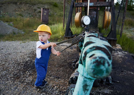 Adorable child in construction helmet using industrial wrench while repairing pipe of oil well pump jack. Cute kid oil worker tightening bolt on pipe. Concept of petroleum industry and childhood.の写真素材