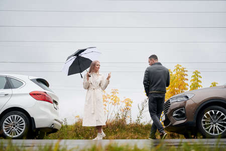 Young couple on the road near cars under cloudy sky with rain, saying goodbye to each other.