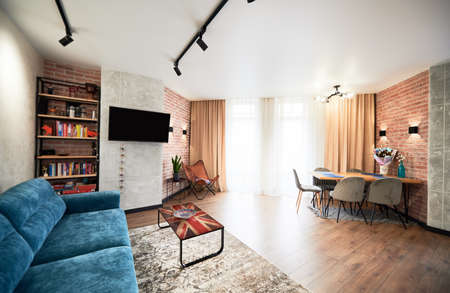 View of interrior of spacious living room with big blue sofa. Bright decoration of appartment with bookshelf, TV, wide window and curpet on wooden floor. Concept of living room design.の写真素材