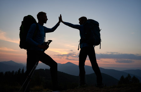 Full length of young man and woman travelers giving high five while hiking together in the mountains during sunset. Concept of travelling, hiking and relationships.の素材 [FY310176076730]