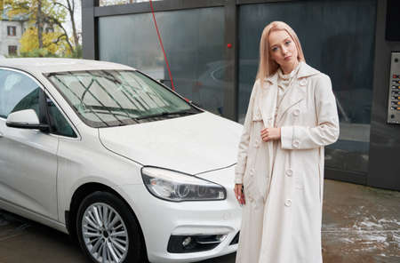 Portrait of young beautiful woman near her white modern car at carwash station outdoor. Female owner...