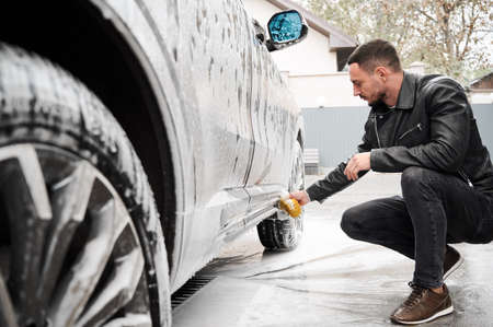Young man washing car on carwash station outdoor. Handsome driver cleaning automobile, using sponge and soap.の写真素材