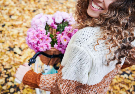 Close up of joyful young woman with curly hair riding bicycle with bouquet of flowers in basket. Female person with beautiful smile holding bike handlebars with pink flowers. Focus on smiling woman.の写真素材