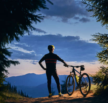 Rear view on a cyclist standing holding his bike, watching beautiful landscape, mountains peaks and sunset, surrounded with spruces. Square picture. Active lifestyle conceptの写真素材